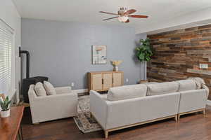 Living room with dark wood finished floors, a ceiling fan, an accent wall, a wood stove, and wooden walls