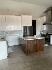 Kitchen with white cabinets, a kitchen island, wood finished floors, stainless steel appliances, and wall chimney range hood