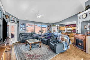 Living room featuring light wood finished floors, a lit fireplace, vaulted ceiling, and ceiling fan