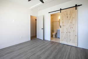 Unfurnished bedroom with a barn door, baseboards, ensuite bath, wood finished floors, and beamed ceiling