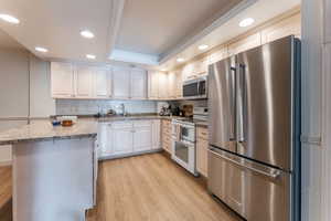 Kitchen with light wood finished floors, appliances with stainless steel finishes, a tray ceiling, backsplash, and recessed lighting