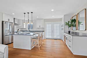 Kitchen with stainless steel fridge, wall chimney exhaust hood, light wood-style flooring, a kitchen breakfast bar, and white cabinetry