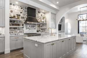 Kitchen featuring arched walkways, decorative backsplash, gray cabinetry, a sink, and wall chimney exhaust hood