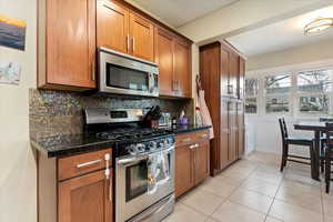 Kitchen with light tile patterned floors, stainless steel appliances, backsplash, brown cabinets, and dark stone counters