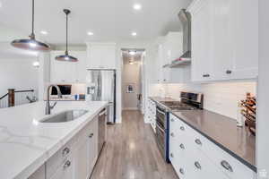 Kitchen with wall chimney exhaust hood, stainless steel appliances, white cabinetry, pendant lighting, and a sink