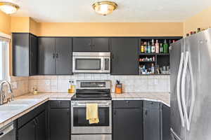 Kitchen with a textured ceiling, a sink, appliances with stainless steel finishes, tile counters, and decorative backsplash