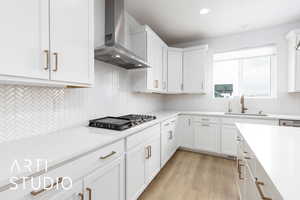 Kitchen with stainless steel gas cooktop, light countertops, white cabinets, a sink, and wall chimney range hood