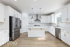 Kitchen with light wood finished floors, wall chimney exhaust hood, a kitchen island, appliances with stainless steel finishes, and a sink