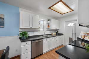 Kitchen with light wood finished floors, dark countertops, white cabinets, a sink, and dishwasher