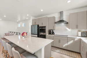 Kitchen featuring double oven, black electric stovetop, gray cabinetry, wall chimney range hood, and quartz countertops