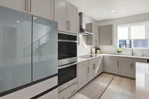 Kitchen with electric stovetop, gray cabinets, decorative backsplash, stainless steel double oven, and wall chimney exhaust hood