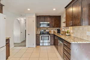 Kitchen with stainless steel appliances, light tile patterned flooring, a sink, dark brown cabinetry, and light stone countertops