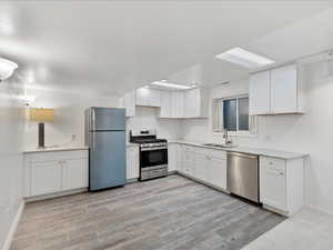 Kitchen with light wood finished floors, appliances with stainless steel finishes, white cabinets, a sink, and baseboards