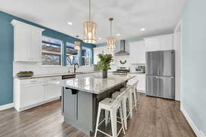 Kitchen featuring white cabinets, wall chimney exhaust hood, appliances with stainless steel finishes, wood finished floors, and a center island