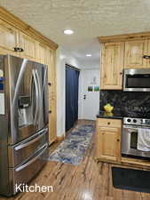 Kitchen with stainless steel appliances, tasteful backsplash, dark wood-type flooring, light brown cabinets, and a textured ceiling