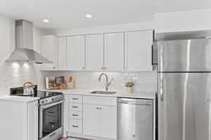 Kitchen featuring stainless steel appliances, light countertops, white cabinets, a sink, and wall chimney range hood