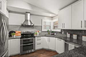 Kitchen with lofted ceiling, stainless steel appliances, a sink, wall chimney range hood, and dark stone counters