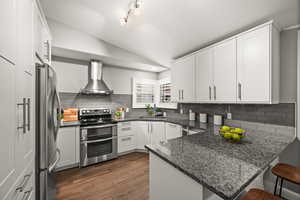 Kitchen with lofted ceiling, a peninsula, a sink, appliances with stainless steel finishes, and wall chimney range hood