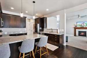 Kitchen featuring stainless steel appliances, a sink, open floor plan, dark brown cabinets, and light stone countertops