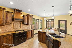 Kitchen featuring decorative backsplash, light stone counters, premium range hood, stainless steel gas cooktop, and a sink