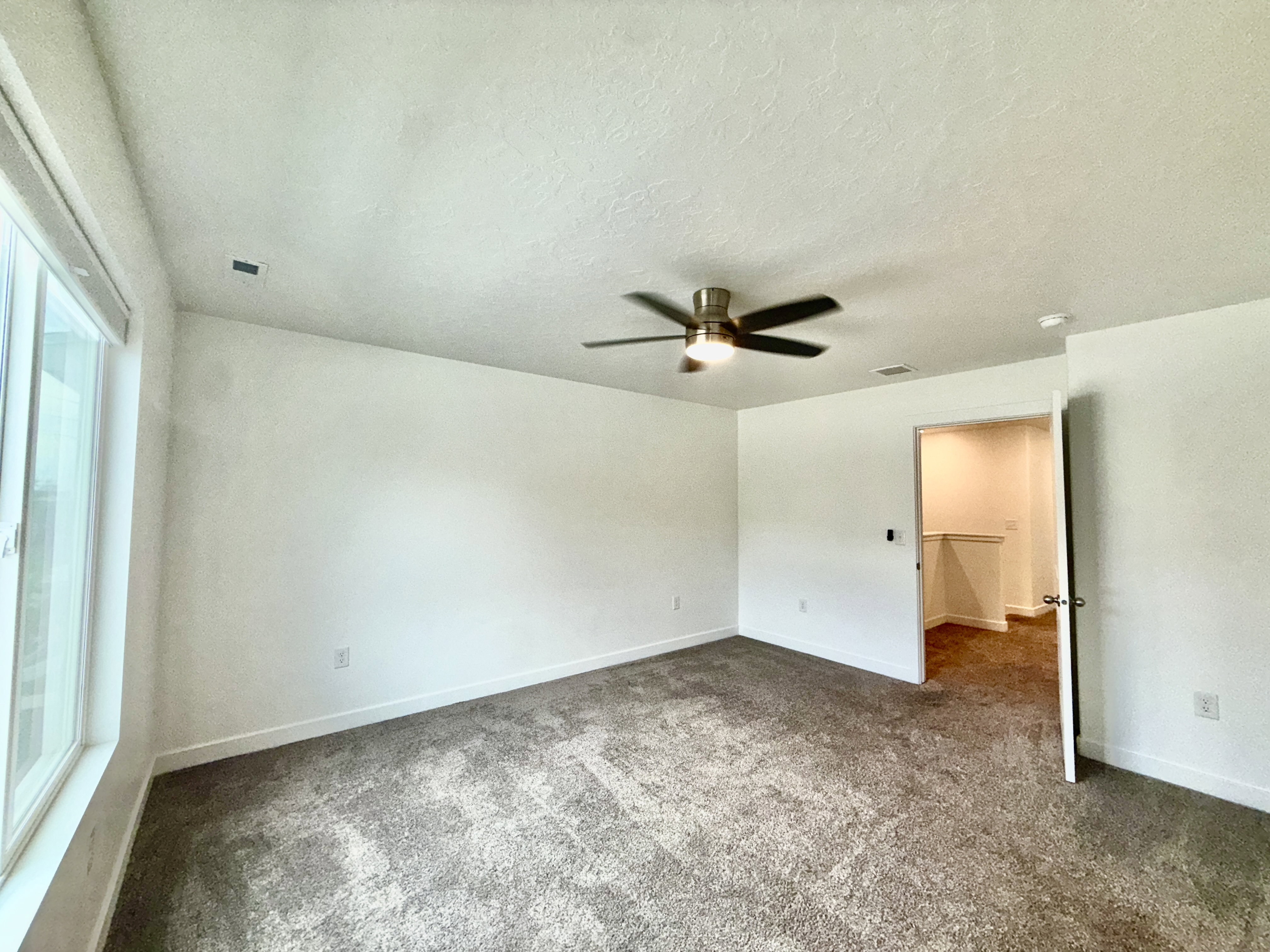 Spacious room with plush gray carpeting, large window, ceiling fan with integrated lighting, and a closet with wood-finish flooring