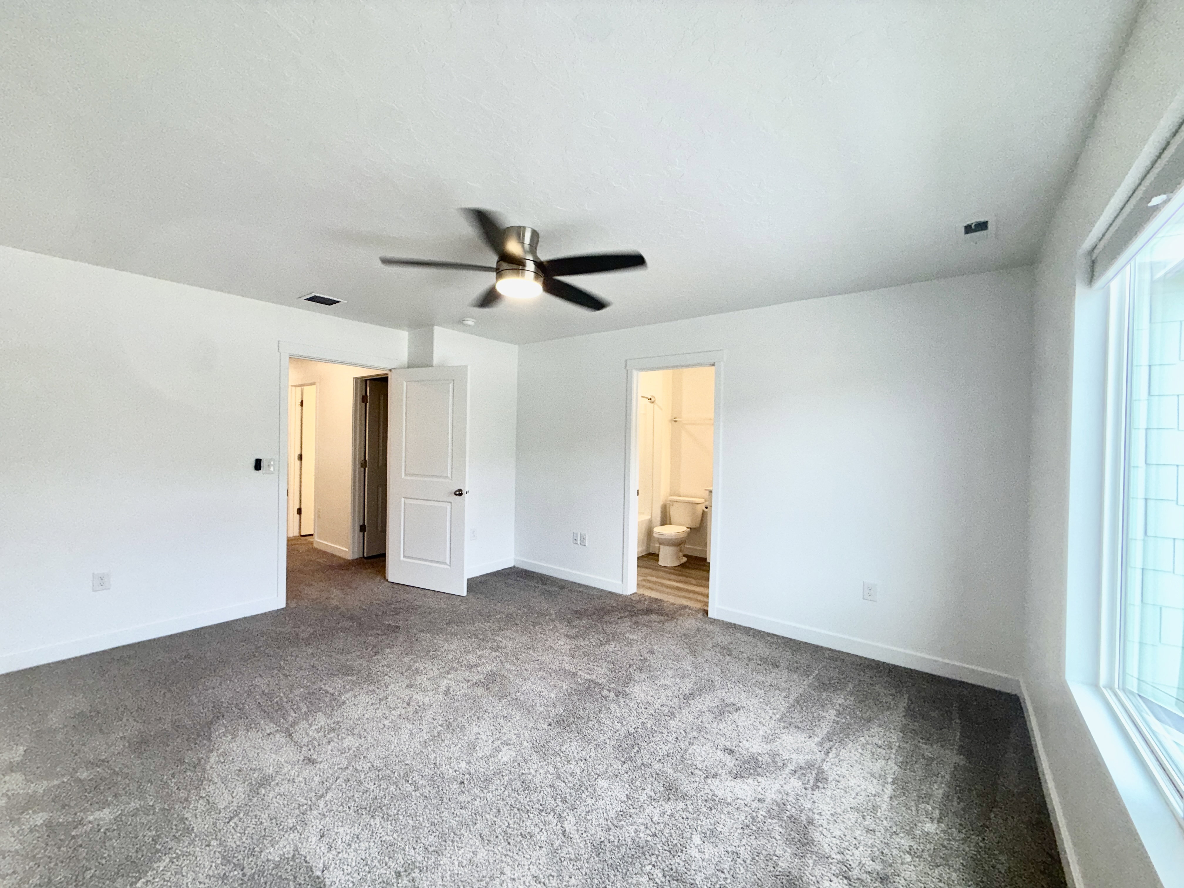 Spacious room featuring neutral carpeting, white walls, a ceiling fan, and a large window