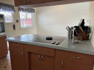 Kitchen with black electric stovetop, light tile patterned floors, stainless steel oven, and brown cabinetry