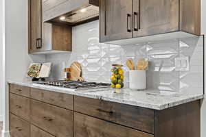 Kitchen featuring premium range hood, stainless steel gas stovetop, decorative backsplash, and light stone countertops
