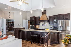 Kitchen featuring dark brown cabinetry, dark wood-style flooring, a spacious island, appliances with stainless steel finishes, and custom range hood