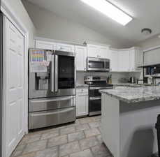 Kitchen featuring white cabinets, decorative backsplash, appliances with stainless steel finishes, light stone countertops, and vaulted ceiling