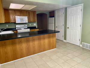 Kitchen with arched walkways, dark countertops, a sink, white appliances, and a peninsula