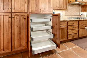 Room details featuring brown cabinets, light stone counters, stainless steel gas stovetop, tasteful backsplash, and under cabinet range hood