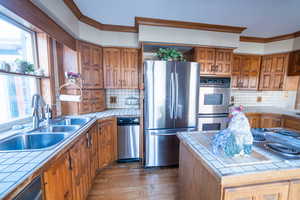 Kitchen featuring crown molding, tile countertops, appliances with stainless steel finishes, brown cabinetry, and a sink