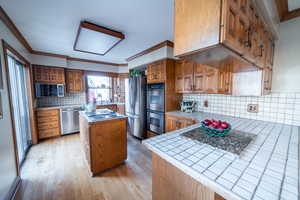 Kitchen featuring stainless steel appliances, tile counters, brown cabinets, and crown molding