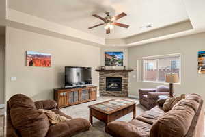 Carpeted living room with baseboards, visible vents, a tray ceiling, a fireplace, and ceiling fan