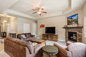 Living area featuring ceiling fan, baseboards, a tray ceiling, light carpet, and a stone fireplace