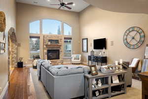 Living area featuring baseboards, light wood-type flooring, a stone fireplace, a high ceiling, and a ceiling fan