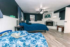 Bedroom featuring a wainscoted wall, a fireplace, wood finished floors, and a ceiling fan