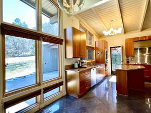 Kitchen with brown cabinets, a notable chandelier, light stone counters, appliances with stainless steel finishes, and vaulted ceiling with beams