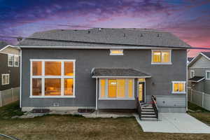 Back of property at dusk with a yard, stucco siding, roof with shingles, and fence