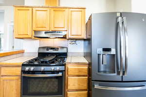 Kitchen featuring appliances with stainless steel finishes, tile counters, light brown cabinets, and under cabinet range hood