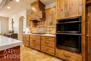 Kitchen with light stone counters, custom exhaust hood, arched walkways, appliances with stainless steel finishes, and hand painted backsplash