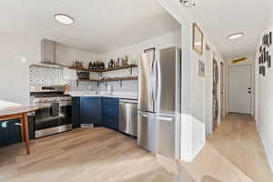 Kitchen with open shelves, blue cabinets, appliances with stainless steel finishes, wall chimney exhaust hood, and light wood-type flooring