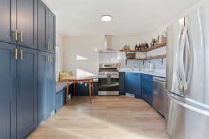 Kitchen with open shelves, light wood-type flooring, appliances with stainless steel finishes, and wall chimney exhaust hood