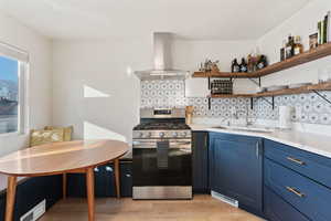 Kitchen with tasteful backsplash, blue cabinetry, stainless steel gas stove, wall chimney exhaust hood, and open shelves