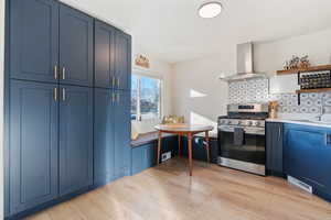 Kitchen featuring light wood-type flooring, blue cabinetry, open shelves, wall chimney exhaust hood, and stainless steel range with gas stovetop