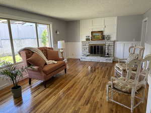Living room featuring wood finished floors, a decorative wall, wainscoting, a stone fireplace, and a textured ceiling