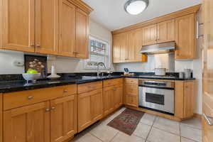 Kitchen featuring under cabinet range hood, stainless steel oven, light tile patterned floors, dark stone countertops, and a sink