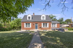 Cape cod house featuring brick siding, a chimney, a front lawn, and a shingled roof