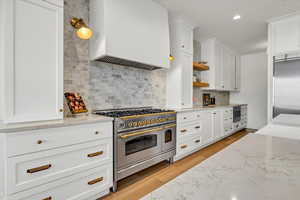 Kitchen featuring light stone countertops, custom exhaust hood, appliances with stainless steel finishes, white cabinetry, and backsplash